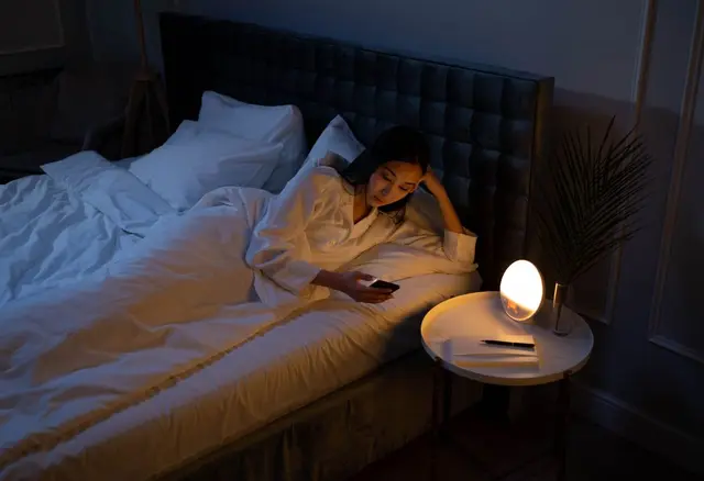 Kobieta w łóżku patrzy w telefon, obok świeci lampka