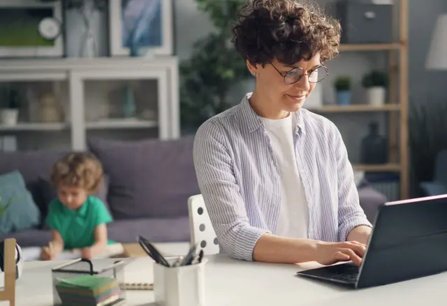 Kobieta pracuje na laptopie, obok bawi się dziecko